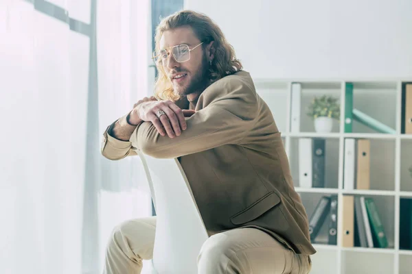 Усміхнений елегантний модний бізнесмен позує на стілець у сонячному світлі в офісі — стокове фото