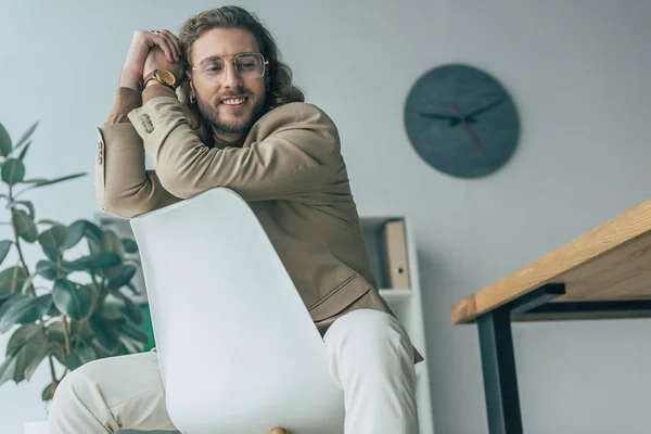 Bajo ángulo vista de sonriente elegante hombre de negocios de moda posando en la silla en la oficina - foto de stock