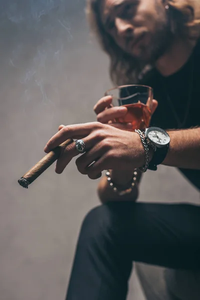 Selektiver Fokus des modischen Geschäftsmannes im schwarzen Outfit, der Zigarre und Whiskey in der Nähe der grauen Wand hält — Stockfoto