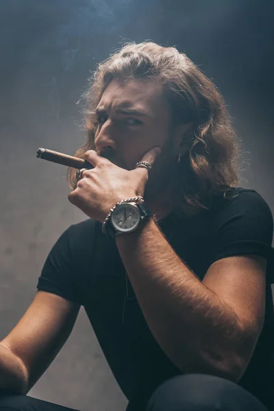 Uomo d'affari alla moda in abito nero fumare sigaro vicino al muro grigio — Foto stock