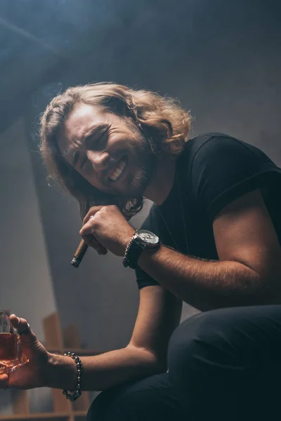 Low angle view of laughing fashionable businessman in black outfit smoking cigar and drinking whiskey — Stock Photo