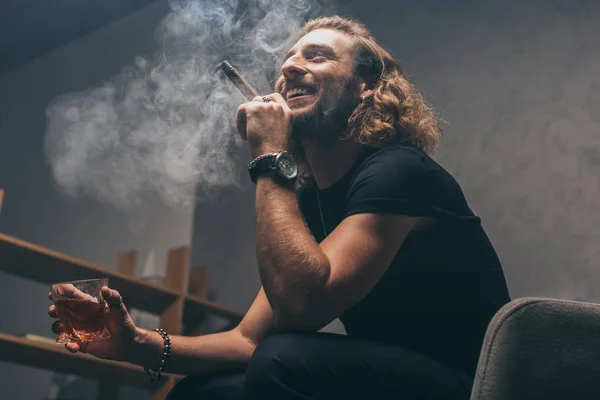 Vista de ángulo bajo de sonriente hombre de negocios de moda en traje negro fumar cigarro y beber whisky - foto de stock