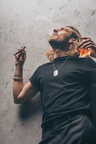 Low angle view of fashionable businessman in black outfit smoking cigar and drinking whiskey near grey wall — Stock Photo