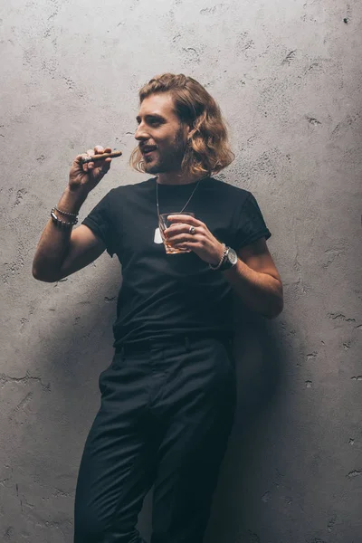 Hombre de negocios de moda en traje negro con cigarro y whisky cerca de la pared gris - foto de stock