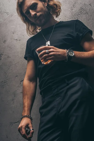 Low angle view of fashionable businessman in black outfit with cigar and whiskey near grey wall — Stock Photo