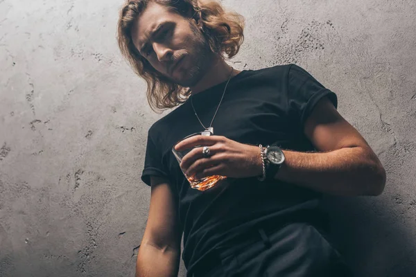 Low angle view of fashionable businessman in black outfit with whiskey near grey wall — Stock Photo