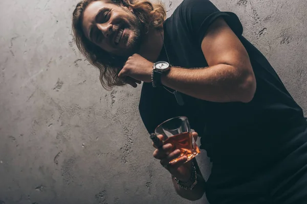 Vista de ángulo bajo del hombre de negocios de moda sonriente en traje negro con cigarro y whisky cerca de la pared gris - foto de stock