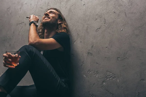 Vista de ángulo bajo de sonriente hombre de negocios de moda en traje negro sentado en el suelo con cigarro y whisky cerca de la pared gris — Stock Photo