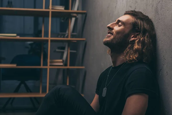 Vista lateral de sonriente hombre de negocios de moda en traje negro sentado cerca de la pared gris - foto de stock