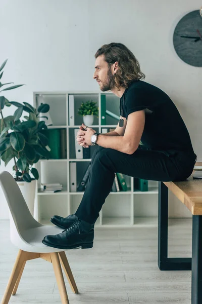 Side view of fashionable casual businessman in total black outfit posing in office on table — Stock Photo