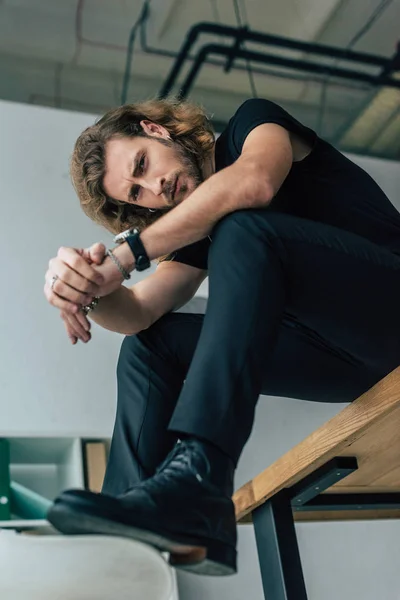 Vista de ángulo bajo del hombre de negocios casual de moda en traje negro total posando en la oficina sobre la mesa - foto de stock