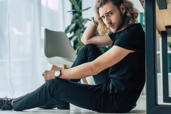 Homem de negócios casual na moda em roupa preta total posando no chão perto da mesa no escritório — Fotografia de Stock