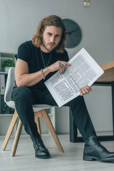 Homem de negócios casual na moda em roupa preta total segurando jornal de negócios na cadeira no escritório — Fotografia de Stock