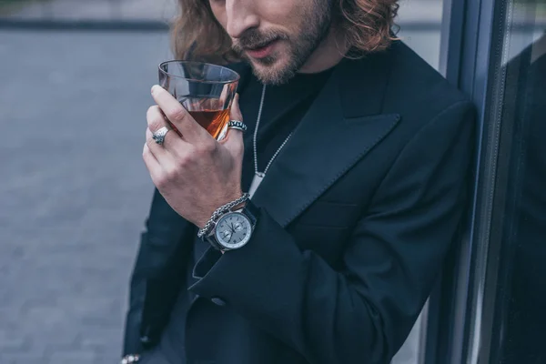 Обрезанный вид на модного бизнесмена в черном костюме, пьющего виски возле офисного здания — стоковое фото