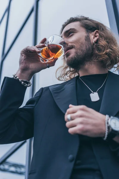 Vista de ángulo bajo del hombre de negocios de moda en traje negro bebiendo whisky cerca del edificio de oficinas - foto de stock