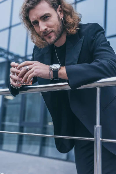 Homem de negócios na moda em terno preto segurando vidro de uísque perto de edifício de escritório e corrimão — Fotografia de Stock