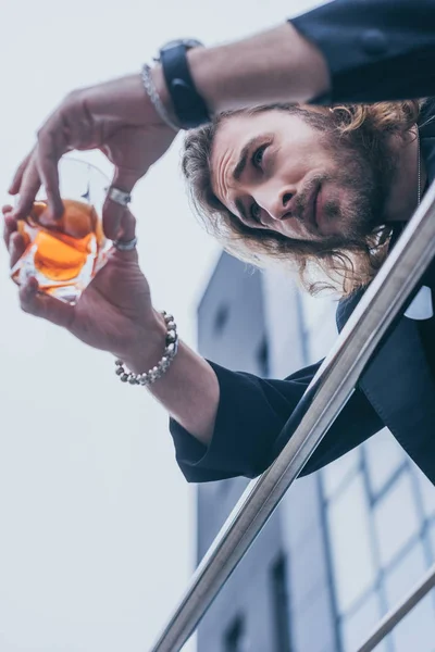Vista de ángulo bajo del hombre de negocios de moda en traje negro sosteniendo vaso de whisky cerca del edificio de oficinas y barandilla - foto de stock