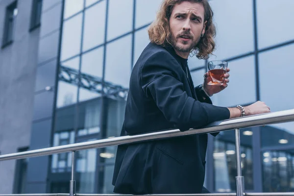 Uomo d'affari alla moda in tuta nera che tiene il bicchiere di whisky vicino a edificio di ufficio e ringhiera — Foto stock