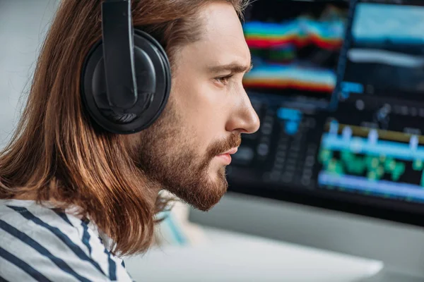 Vue latérale de l'éditeur barbu écoutant de la musique dans les écouteurs près du moniteur d'ordinateur — Photo de stock