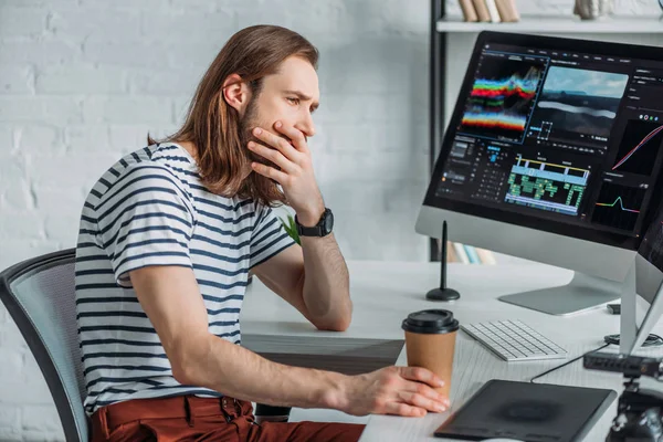 Editor holding paper cup while covering mouth and looking at computer monitor — Stock Photo