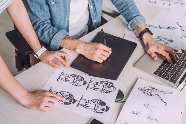 Vista recortada del ilustrador apuntando con el dedo al boceto de dibujos animados cerca de compañero de trabajo - foto de stock