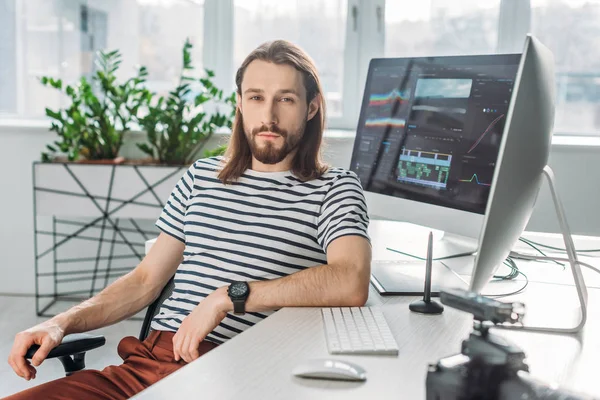Избирательный фокус красивого бородатого редактора, сидящего рядом с мониторами компьютера — стоковое фото