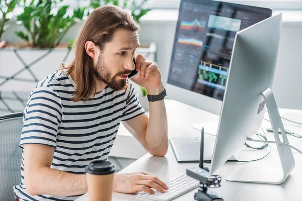 Bearded art editor talking on smartphone near computer monitors — Stock Photo