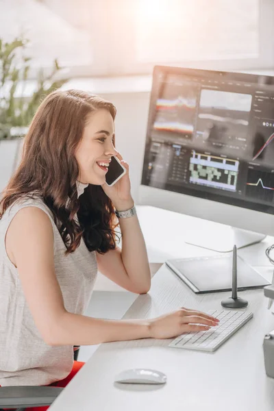 Seitenansicht eines glücklichen Redakteurs, der am Smartphone in der Nähe des Computermonitors spricht — Stockfoto