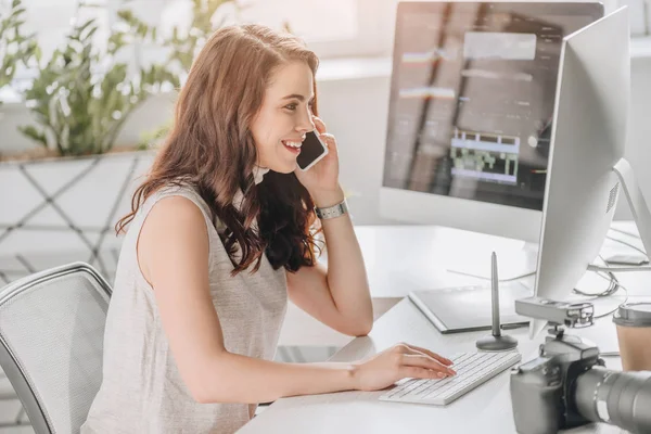 Seitenansicht des gut gelaunten Redakteurs, der auf dem Smartphone neben dem Computermonitor spricht — Stockfoto