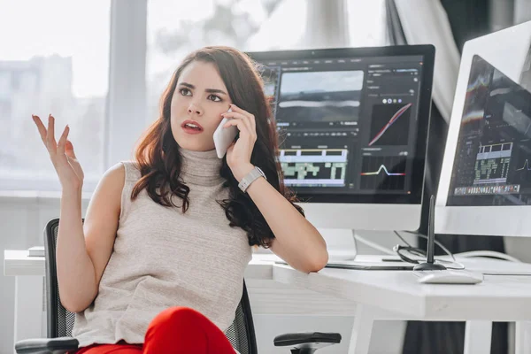 Editor disgustado hablando en el teléfono inteligente y el gesto cerca de los monitores de ordenador - foto de stock