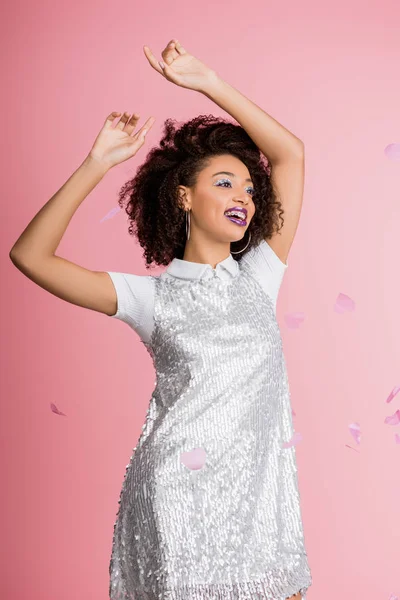 Séduisante fille afro-américaine heureuse avec bretelles dentaires, avec fards à paupières à paillettes argentées et lèvres violettes dansant en robe paillettes, isolée sur rose avec confettis — Photo de stock