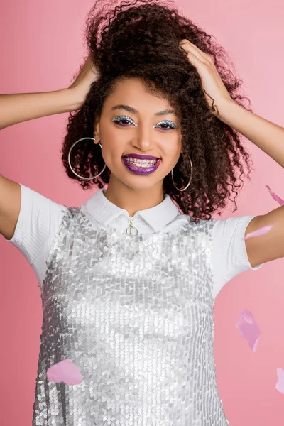 Cheerful african american girl with dental braces, with silver glitter eyeshadows and purple lips wearing paillettes dress, isolated on pink with confetti — Stock Photo