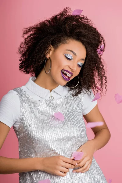 Positive african american girl with dental braces, with silver glitter eyeshadows and purple lips wearing paillettes dress, isolated on pink with confetti — Stock Photo
