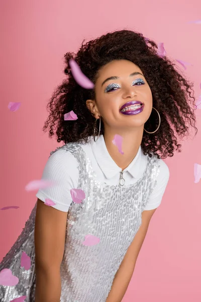 Funny african american girl with dental braces, with silver glitter eyeshadows and purple lips wearing paillettes dress, isolated on pink with confetti — Stock Photo