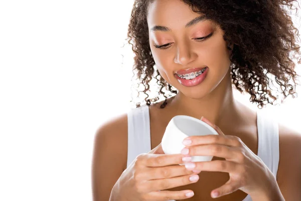 Niña afroamericana sonriente sosteniendo contenedor de plástico con crema facial, aislado en blanco - foto de stock