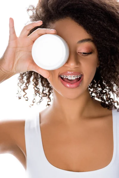 Menina americana africana alegre com aparelho dentário segurando recipiente de plástico com creme facial, isolado em branco — Fotografia de Stock