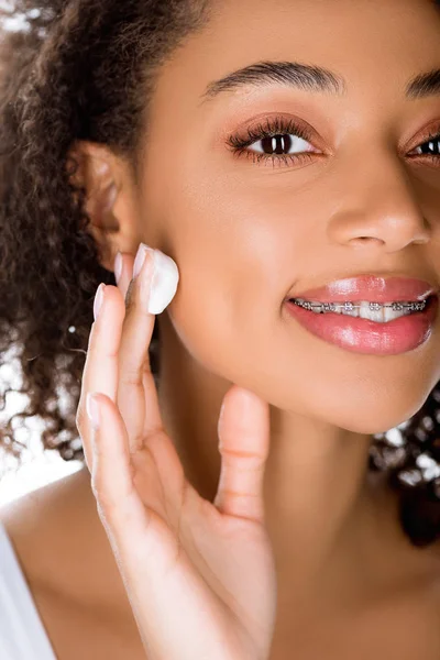 Felice ragazza afroamericana con bretelle dentali applicare crema idratante, isolato su bianco — Foto stock