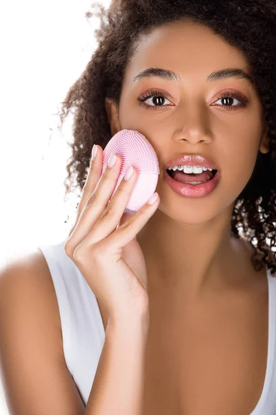 Sorpreso ragazza afro-americana con bretelle utilizzando spazzola viso detergente in silicone, isolato su bianco — Foto stock