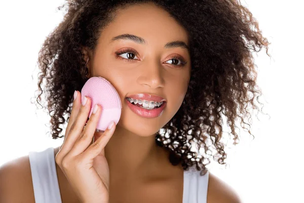 Atractiva chica afroamericana feliz con frenos usando cepillo facial limpiador de silicona, aislado en blanco - foto de stock