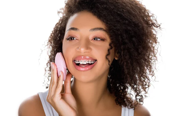 Souriant afro-américaine fille avec des appareils dentaires à l'aide de silicone nettoyage brosse faciale, isolé sur blanc — Photo de stock