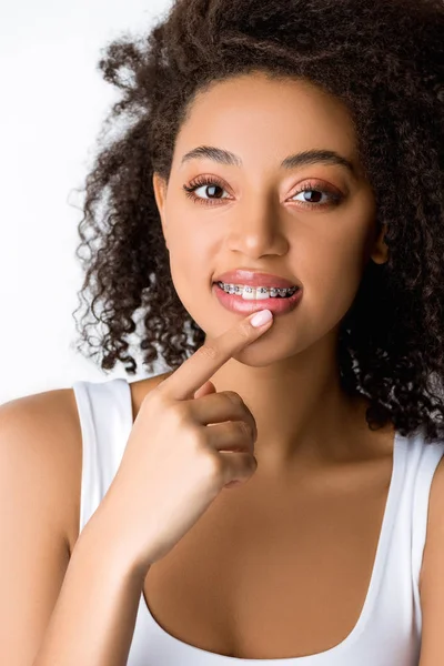 Пухнаста усміхнена афроамериканська дівчина з зубними брекетами, торкаючись губ, ізольована на сірому — стокове фото