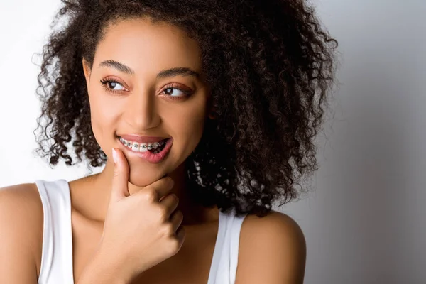 Pensativo sorrindo menina afro-americana com aparelho dental tocando lábio, isolado em cinza — Fotografia de Stock