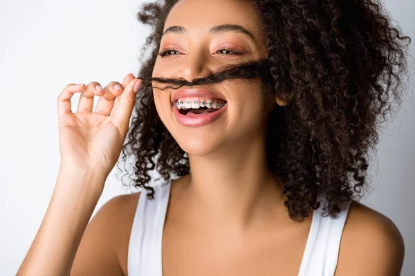 Rindo menina afro-americana com aparelho dentário fazendo bigode falso do cabelo, isolado em cinza — Fotografia de Stock