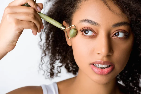 Belle frisée afro-américaine fille à l'aide de jade rouleau facial, isolé sur gris — Photo de stock