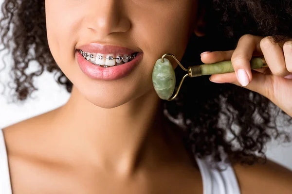 Обрізаний вид усміхненої афроамериканської дівчини з зубними брекетами з використанням натурального масажного ролика, ізольований на сірому — стокове фото