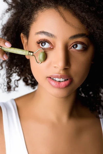 Beautiful african american girl using jade facial roller, isolated on grey — Stock Photo