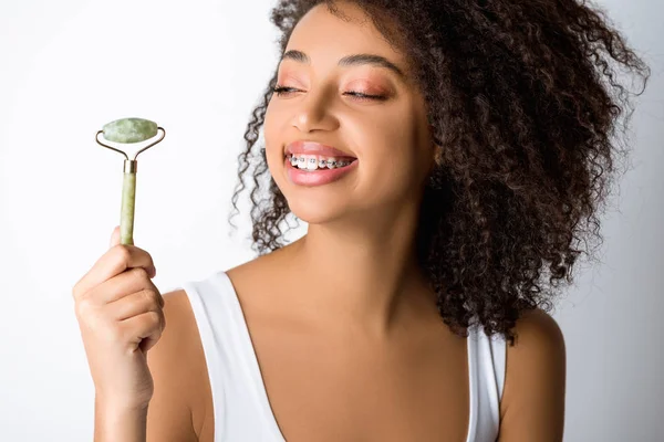 Menina afro-americana feliz com aparelho dentário usando rolo de massagem natural, isolado em cinza — Fotografia de Stock