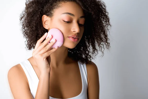 Curly african american girl using silicone cleansing facial brush, isolated on grey — Stock Photo