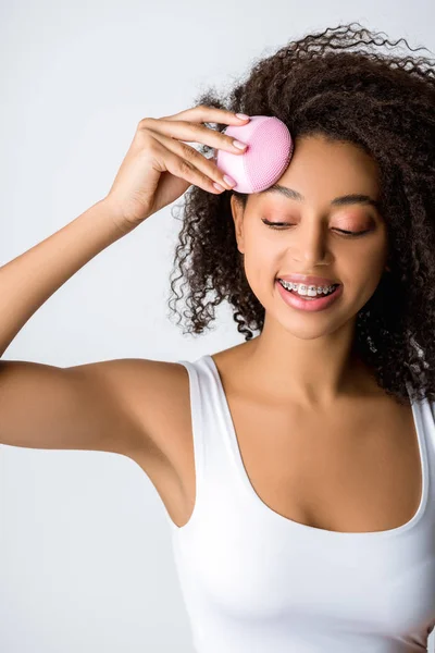 Smiling african american woman with braces using silicone cleansing facial brush, isolated on grey — Stock Photo