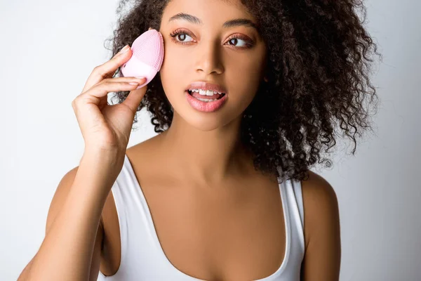 Jeune femme afro-américaine utilisant une brosse faciale nettoyante en silicone, isolée sur gris — Photo de stock